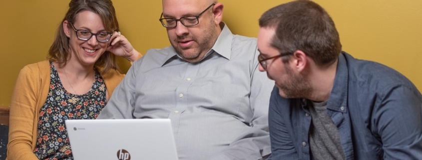 A woman and two men sitting on a bench looking at a laptop in front of a yellow wall.