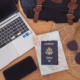 Flat lay of an open laptop, camera, phone, passport, map, travel bag, sun hat, and sunglasses on a brown table.