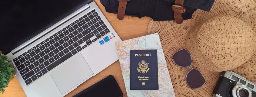 Flat lay of an open laptop, camera, phone, passport, map, travel bag, sun hat, and sunglasses on a brown table.