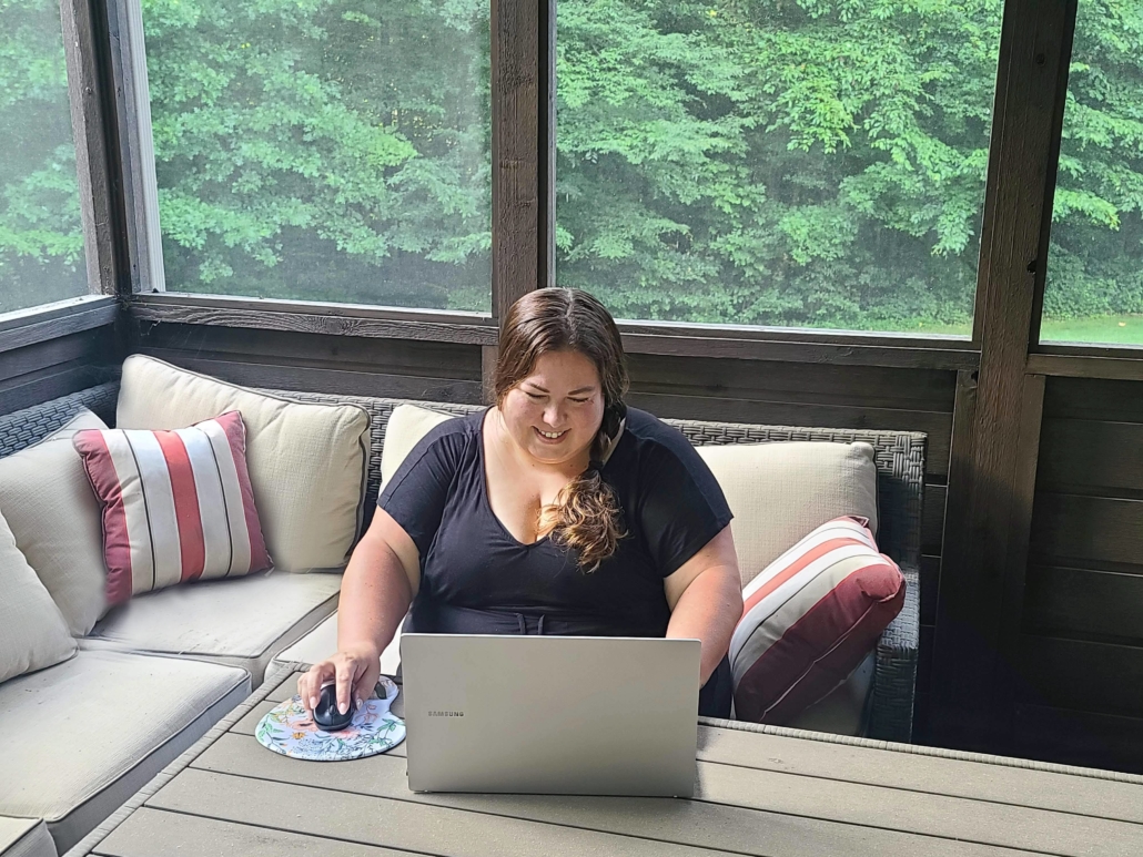 Kelly sitting on a couch in a screened-in porch with a laptop and mouse in front of her. Kelly is wearing black with her hair down. The couch has striped decorative pillows and the background has lush green trees just outside the windows.